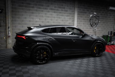Gösterişli, siyah bir Lamborghini Urus Performante kapalı bir yerde, kareli bir zeminde park halinde. Dekor, içinde aslan amblemi olan beton blok bir duvar içeriyor..