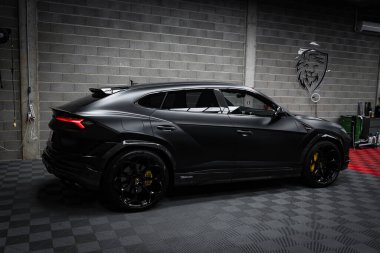 A sleek black Lamborghini Urus Performante is parked indoors on a checkered floor. The setting includes a concrete block wall with a lion emblem, highlighting luxury. clipart