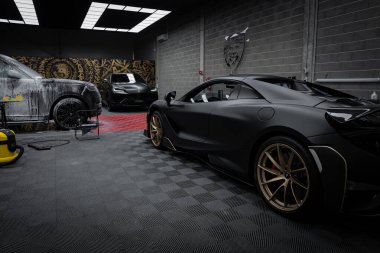 Altın jantlı siyah bir McLaren 720S modern bir garajda kareli zemine park edilmiştir. Arka planda bir Lamborghini Urus var, kısmen sabunla kaplı..