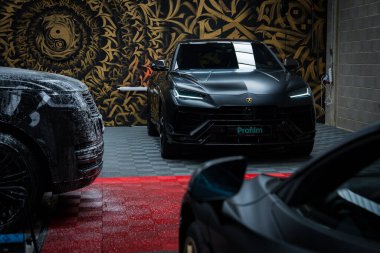 Parlak, siyah bir Lamborghini Urus Performante, dönen bir duvar resmiyle bir garajda sergileniyor. Kırmızı ve gri fayanslı zemin ve diğer lüks arabaların parçaları görünür..