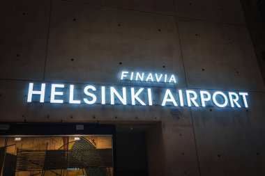 Görüntü, Finavia Helsinki Havalimanı 'nın koyu arkaplan üzerinde, modern havaalanı tasarımını ön plana çıkaran, sanatsal bir duvar resmi olan parlak ışıklı bir tabela gösteriyor..