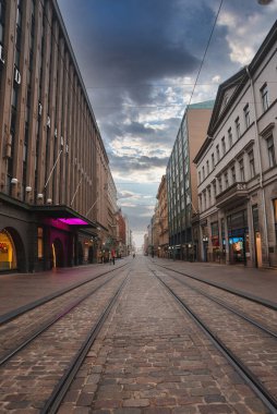Helsinki, Finlandiya 'da tramvay rayları, tarihi binalar ve parçalı bulutlu bir gökyüzünün bulunduğu kaldırım taşı caddesi manzarası sakin bir şehir manzarası yaratıyor..