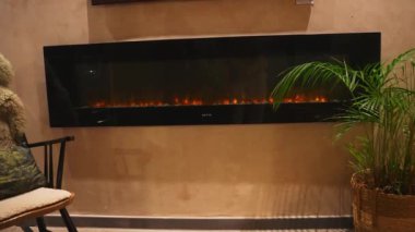 Konforlu bir yaşam alanı, bir duvara monte edilmiş elektrikli şömine, dekoratif yastıklı peluş bir sandalye ve yeşillik ekleyen bir saksı bitkisine sahiptir..