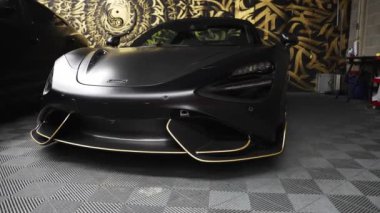 McLaren 720S garajda sergileniyor, mat siyah kaplamasını, altın aksanını ve keskin farlarını kareli zemine vurguluyor..