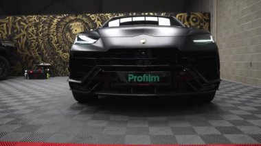 Video, Lamborghini Urus 'u kapalı bir garajda siyah bir kaplamayla çekiyor, arka planda endüstriyel duvarlar ve benzersiz bir sanatsal duvar resmi var..