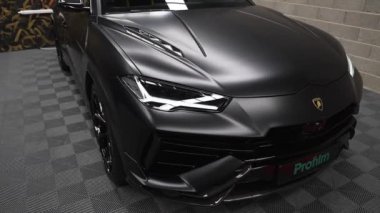 Video Lamborghini Urus 'un mat siyah dış görünüşünü yakalıyor, amblem ve açısal farlara odaklanıyor, kareli zemine ve dekoratif duvara yaslanıyor..