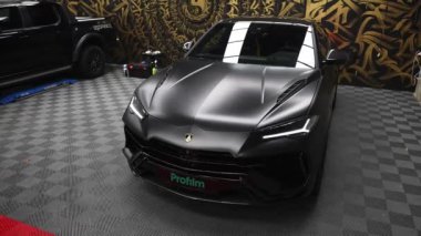 Video sergi salonunda siyah bir Lamborghini Urus sergiliyor, açısal tasarımını ve şablonlu duvarlara karşı lüks detaylarını vurguluyor..