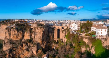 Aerial view of Ronda, Spain, at sunset with Puente Nuevo bridge over a deep gorge, surrounded by cliffs, greenery, and whitewashed Andalusian buildings. clipart