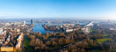 Morning light illuminates Riga's skyline, featuring the Daugava River, National Library, Vansu Bridge, and Ferris wheel in Victory Park. clipart