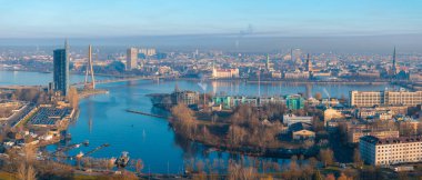 Aerial view of Riga, Latvia, featuring the Daugava River, Vansu Bridge, and the Ferris wheel in Victory Park. Morning light highlights the city's skyline. clipart