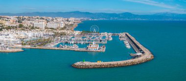 The image captures Benalmadena's marina with yachts, a Ferris wheel, and a curved breakwater. Distant mountains and town buildings are visible under a clear sky. clipart