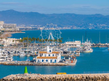 Benalmadena, İspanya 'nın canlı marinasında bir dönme dolap, yatlar ve mavi aksanlı beyaz bir bina bulunur. Palmiye ağaçları ve dağlar kıyı boyunca uzanır..