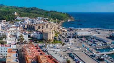 Tarifa, Spain, features Guzman Castle, whitewashed buildings, and the Strait of Gibraltar. The marina is active, surrounded by green hills and blue waters. clipart