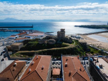 İspanya, Tarifa 'nın havadan görünüşü, kıyıya yakın Guzman Kalesi' ni vurguluyor. Sahnede kumlu bir sahil, dalgakıranlar, uçurtma sörfçüleri ve kırmızı kiremitli çatılar yer alıyor..