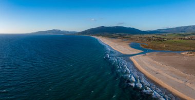 Tarifa, İspanya 'nın havadan görünüşü, kumlu bir plaj, Atlantik Okyanusu ve yeşil tepeler. Kıyı şeridi yumuşak dalgalarla berrak bir gökyüzü altında yavaşça kıvrılır..