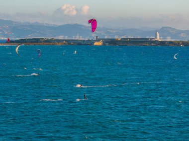 İspanya, Tarifa yakınlarındaki canlı mavi sulardaki uçurtma sörfçülerinin hava perspektifi. Sahilde bir deniz feneri duruyor, arka planda rüzgar türbinleri ve dağlar var..