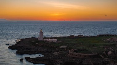 The image shows a sunset over Tarifa, Spain, featuring a lighthouse on a rocky outcrop. Historical ruins and distant ships are visible along the expansive ocean. clipart