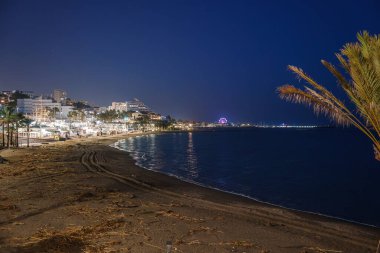 Benalmadena plajının sakin bir gece sahnesi, aydınlatılmış binalar, palmiye ağaçları ve sakin suları yansıtan uzak bir dönme dolap..