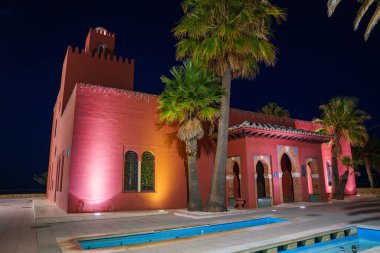 Bil Bil Castle in Benalmadena, Spain, features Moorish architecture with red stucco walls and arched windows, illuminated at night with palm trees and a pool. clipart