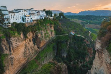 Ronda, İspanya, sarp bir uçurumun tepesinde beyazlatılmış binalarla birlikte. Günbatımı şehrin üzerine ılık bir ışık saçıyor ve kayalıklarda yemyeşil bir alan var..
