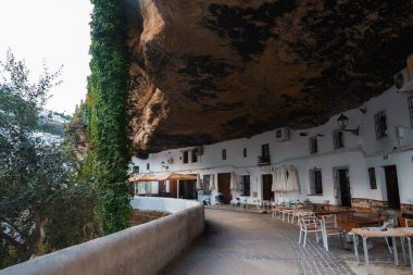 Whitewashed buildings are built into the rock face, with a natural rock canopy above. Outdoor seating and greenery enhance the unique urban landscape. clipart