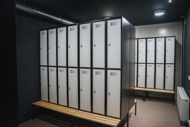 İyi aydınlatılmış bir soyunma odasında ahşap bankların üzerine monte edilmiş havalandırma yuvaları ve güvenli kilitleri olan parlak metalik dolaplar bulunuyor. Karanlık duvarlar ve görünür kanallar endüstriyel bir dokunuş ekliyor..
