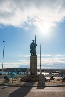 İspanya 'nın Tarifa kentindeki bir heykel, tabanında bir çapa bulunan uzun bir nesneyi tutuyor. Arka planda tekneleri ve açık gökyüzü olan bir marina yer alıyor..