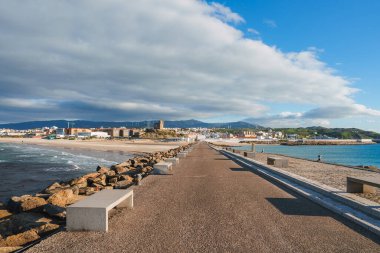 İspanya 'nın Tarifa kentinde uzun bir gezinti güvertesi, kayalık kıyılar ve kumlu bir plaj bulunur. Tanınmış bir kule ve yuvarlanan tepeler parçalı bulutlu bir gökyüzünün altında görülebilir..