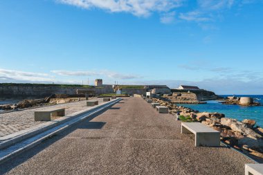 Tarifa, Spain features a stone pathway leading to a historic fortification, possibly the Castle of Guzman, with rocky shores and the Atlantic Ocean nearby. clipart