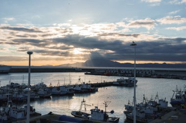 Tarifa limanındaki balıkçı tekneleri, İspanya 'da, bulutların arasından güneş ışınları geçen bir gökyüzünün altında. Bir dalgakıran ve kemerler rıhtımda, uzaktaki dağlarla..
