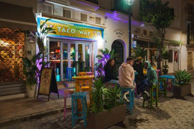 İspanya 'nın Tarifa kentinde Taco Yolu adında renkli bir taco restoranında parlak neon ışıklar, bereketli saksı bitkileri ve çok renkli taburelerde oturan insanlar yer alıyor..