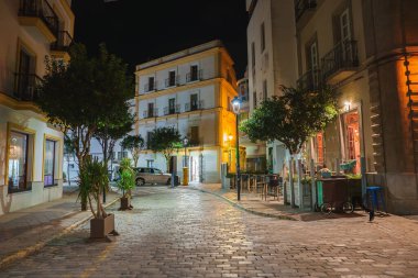 İspanya 'nın Tarifa kentinde sakin bir caddede kaldırım taşları, balkonlu antika binalar ve sıcak ışıklandırma yer alıyor. Ağaçlar ve kafe masaları dingin atmosfere katkı sağlar..