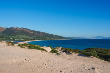Geniş kum tepeleri İspanya 'nın Tarifa kentindeki derin mavi denize çıkar. Uzakta Afrika kıyıları ile rüzgar türbinleri ile yuvarlanan tepeler görünür.