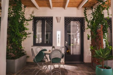 Bahçe alanında metal sandalyeli ve yeşil yastıklı yuvarlak bir masa bulunur. Kemerli pencereler ve kafesler duvarları süslüyor, yeşillik tırmanıyor..