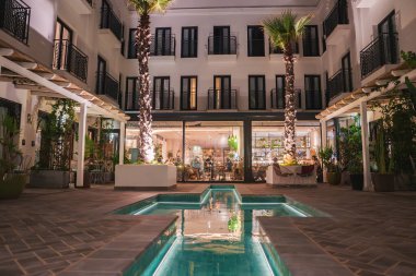 Marakeş tarzında bir avluda geometrik bir havuz, uzun palmiye ağaçları ve yemyeşil yeşillikler bulunur. Demir parmaklıklı balkonlar manzaraya bakıyor..