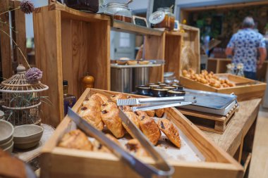 Kahvaltı büfesinde maşalı ahşap tepsiler, kavanozlu kırsal raflar ve davetkar bir atmosfer yaratan günlük kıyafetler içindeki bir insan bulunur..