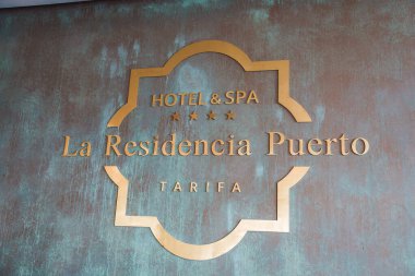 Görüntü, Tarifa 'da bir otel ve spa olan La Residencia Puerto' nun üç yıldızlı, dokulu arka planda altın harfli tabelalarını gösteriyor..