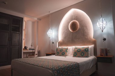 A hotel room features a unique arched headboard, ambient lighting, and Moroccan decor. Intricate pillows, a bed runner, and hanging lanterns enhance the design. clipart