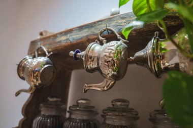 Gümüş çaydanlıklar ahşap bir rafta asılı, altında da kırık cam kavanozlar var. Yeşil bir bitki sağı çerçeveler ve Marakeş sahnesine doğal enerji katar..