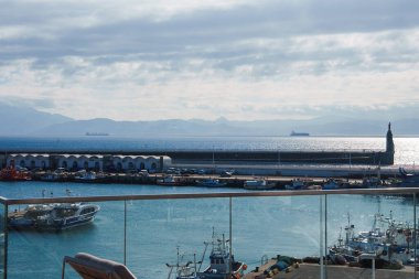 A harbor scene with a breakwater and monument, vibrant blue water, docked boats, and distant mountains under a partly cloudy sky, viewed from a terrace. clipart