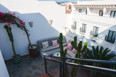 Çatı terasında çiçek yastıkları, canlı begonvilleri ve uzun kaktüsleri olan gri bir kanepe bulunur. Fas kavisli pencereli bir binaya bakar..