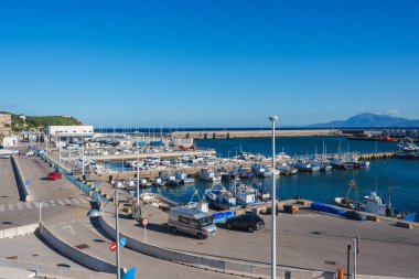 Tangier 'in marinası boyunca sayısız balıkçı teknesi rıhtıma yanaşıyor, açık mavi bir gökyüzüne ve uzak dağlara karşı kuruluyor, yakınlarda iyi korunmuş bir yol var..