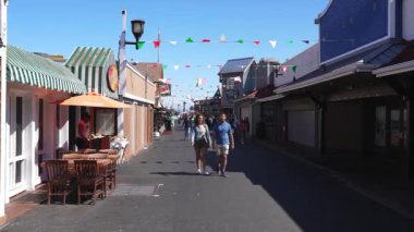 İnsanlar Balıkçı İskelesi 'ndeki dükkan ve restoranlarla dolu kalabalık bir patika boyunca yürüyor. Renkli bayraklar ve açık hava koltukları canlı atmosferi artırır.