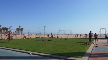 İnsanlar Venedik Plajı, Los Angeles 'ta spor ve egzersiz yapıyor. Açık mavi gökyüzünün altında salıncaklar ve okyanus arkaplanı var..