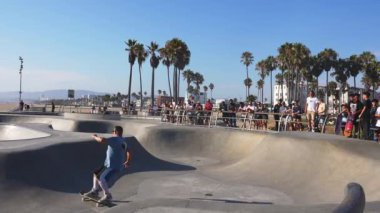 Her yaştan patenciler, Los Angeles 'taki Venice Beach Paten Parkı' nda palmiye ağaçları ve arka planda açık mavi gökyüzünün altında plaj manzarası olan numaralar sergiliyor..