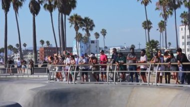 Los Angeles 'taki Venice Beach kaykay parkında palmiye ağaçları, seyirciler ve sahile bakan ikonik mimari ile çevrili patenciler numaralar yapıyor..