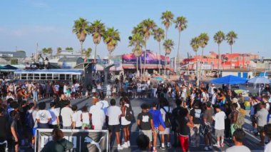 Canlı bir kalabalık Venedik Plajı basketbol sahasında toplanıyor. Palmiye ağaçları, renkli binalar ve Los Angeles 'ta açık mavi gökyüzünün altında bir otobüs..