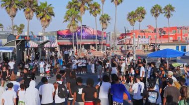 Venedik Plajı, Los Angeles 'ta canlı bir basketbol maçı. Çeşitli izleyiciler, ikonik palmiye ağaçları, renkli duvar resimleri ve görünür bir Muscle Beach tabelası..