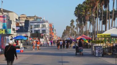 Los Angeles 'taki Venice Beach' te açık mavi gökyüzü altında çeşitli kalabalıklar bulunan hareketli bir sahil yolu, palmiye ağaçları, sokak satıcıları ve duvar resimleri bulunuyor..