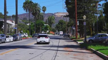 Los Angeles 'ta bir sokakta, parlak yeşil gökyüzü altında palmiye ağaçları ve park edilmiş arabalarla dolu ikonik bir Hollywood tabelası var..